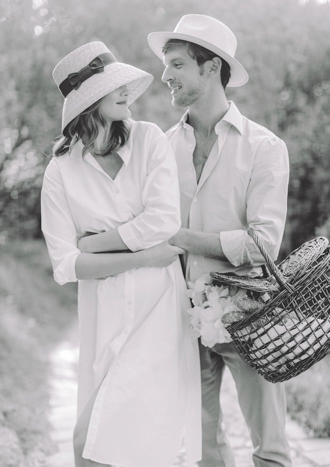 A couple, both wearing hats, smiling at each other and holding a picnic basket. 