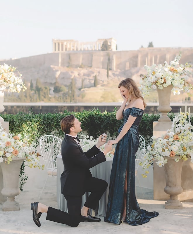 A man on one knee proposing to a woman in a blue shimmering gown. 