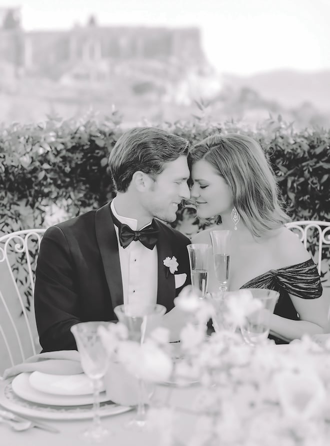 A bride and groom smiling with their foreheads together holding champagne glasses at the table. 