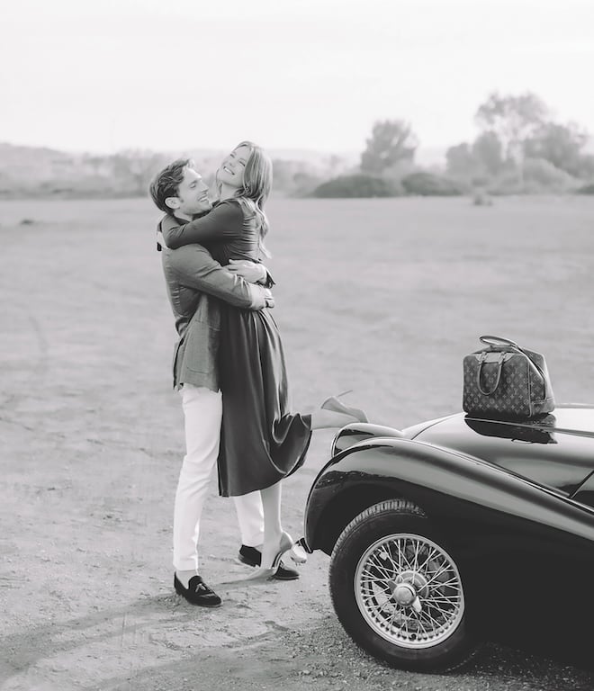 A man hugging and holding his fiancee next to a black vintage car with a Louis Vuitton travel bag on the hood. 