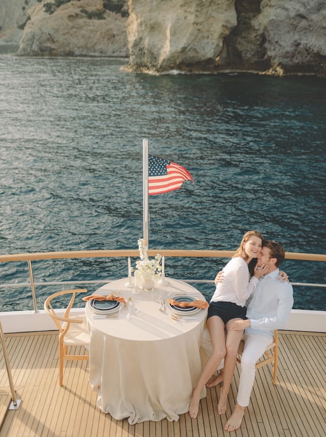 A couple sitting and hugging on a boat next to a table set for two. 