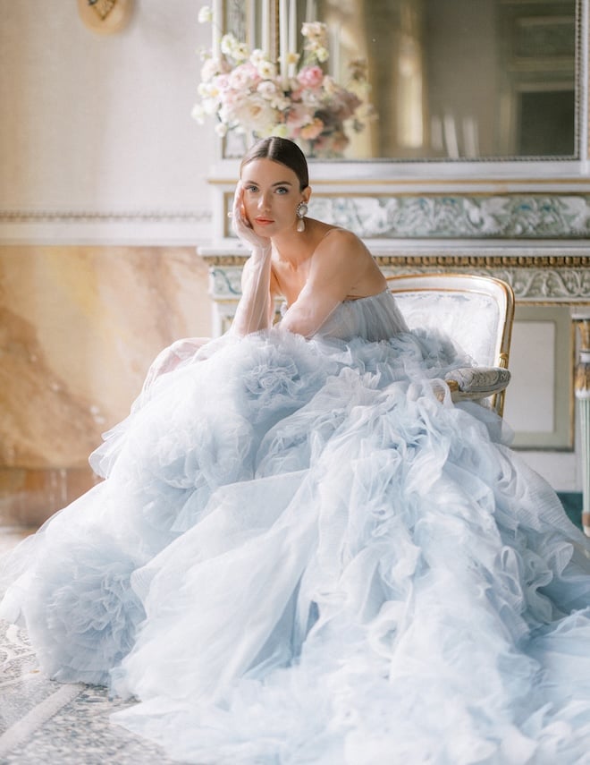 A bride wearing a blue wedding dress sits on a chair. 