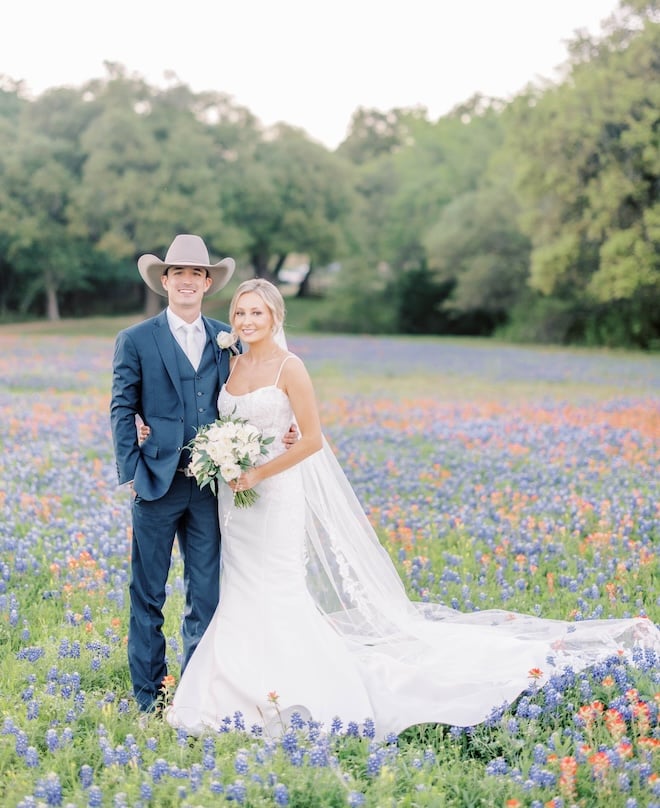 The bride and groom smile standing in a field of wild flowers on their family ranch. 