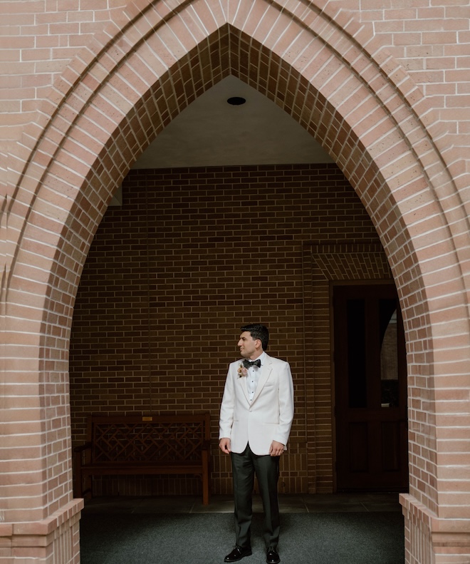 The groom wearing a white suit jacket looking to his side. 