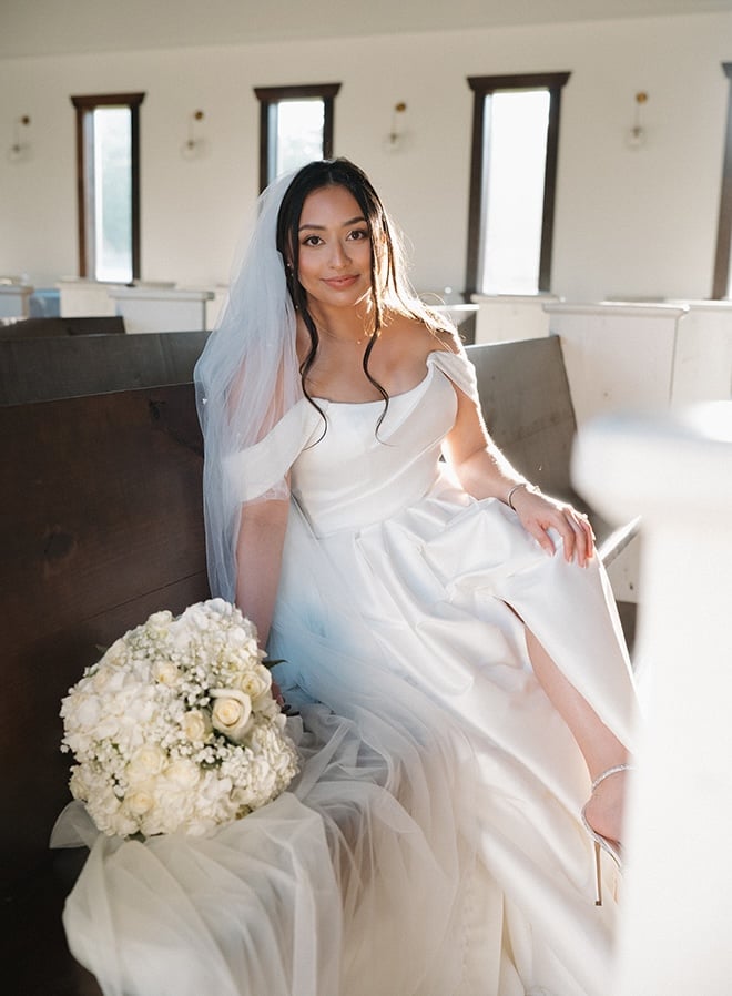 The bride sits on a pew in the chapel wearing her wedding dress. 