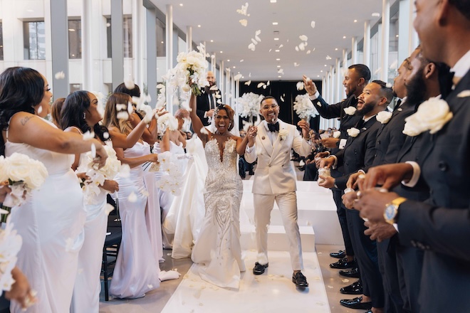 A bride and groom holding hands walking while the wedding party throws flower petals,