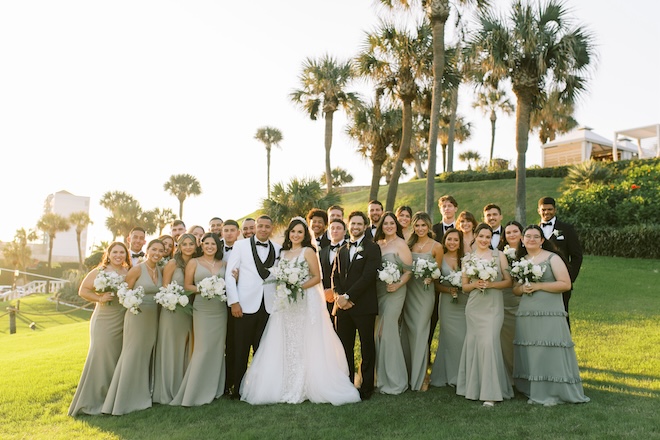 A wedding party in green and black on the lawn of the San Luis Resort with palm trees and landscape behind them. 