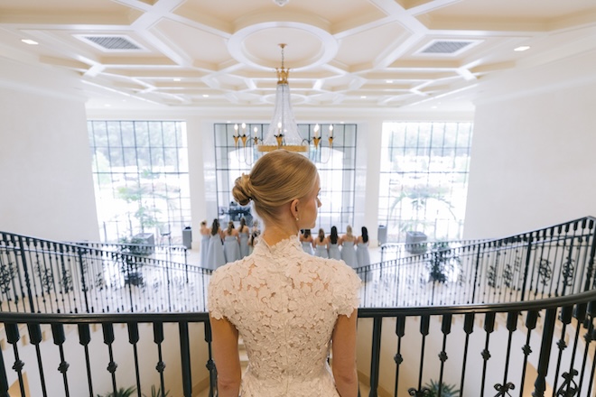 The bride at the top of a staircase with her bridesmaids below waiting to see her dress. 