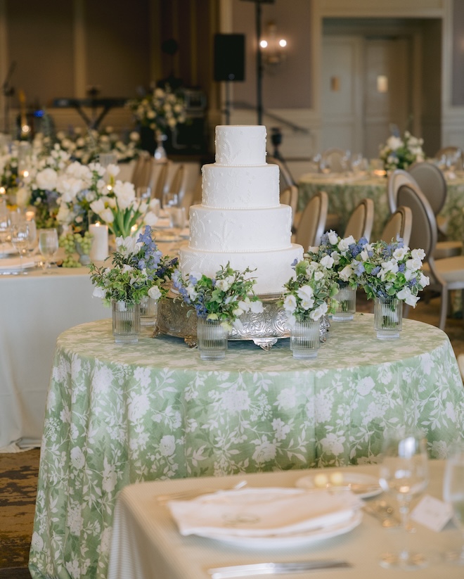 The four-tier white cake with florals around the bottom sitting on a round table in a green floral print tablecloth. 