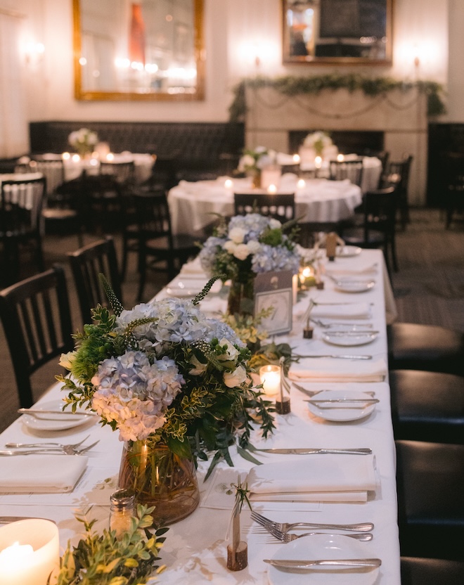 A long table at Ouisie's Table decorated with white linens and blue and green floral arrangements. 