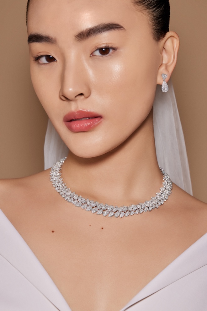 A bride wearing a veil with her hair slicked back wearing diamond drop earrings and a diamond necklace. 