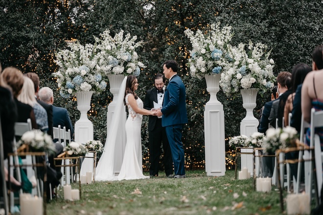 The bride and groom at the altar holding hands surrounded by potted floral arrangements with blue and white florals. 