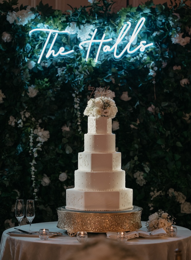 5-tier white cake with flowers on the top and a lit-up blue sign above that reads "The Halls". 