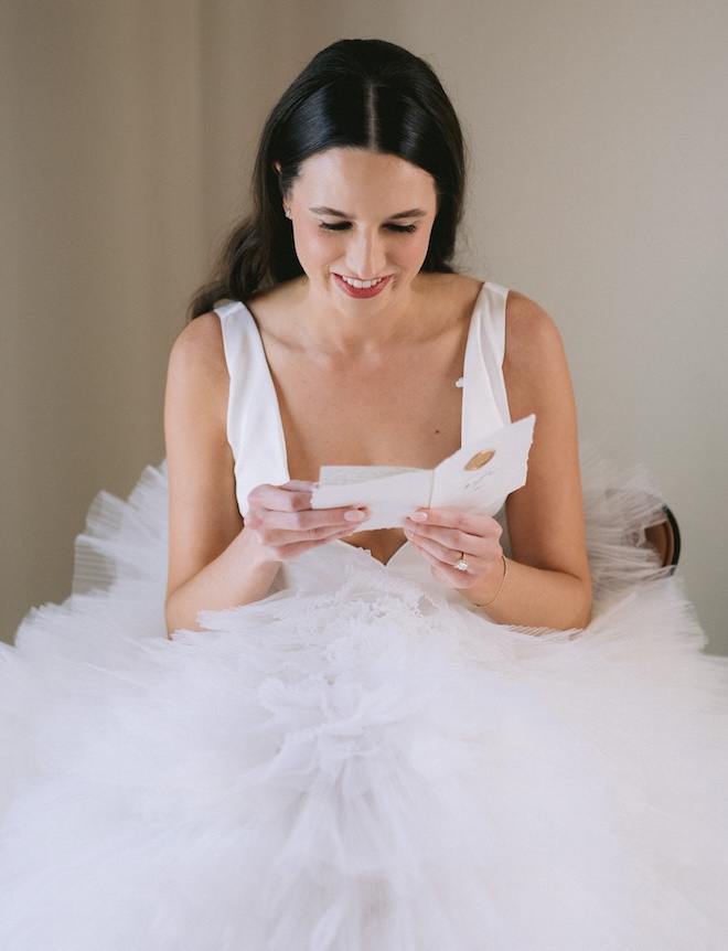 The bride sits in a chair while reading a handwritten note from the groom. 