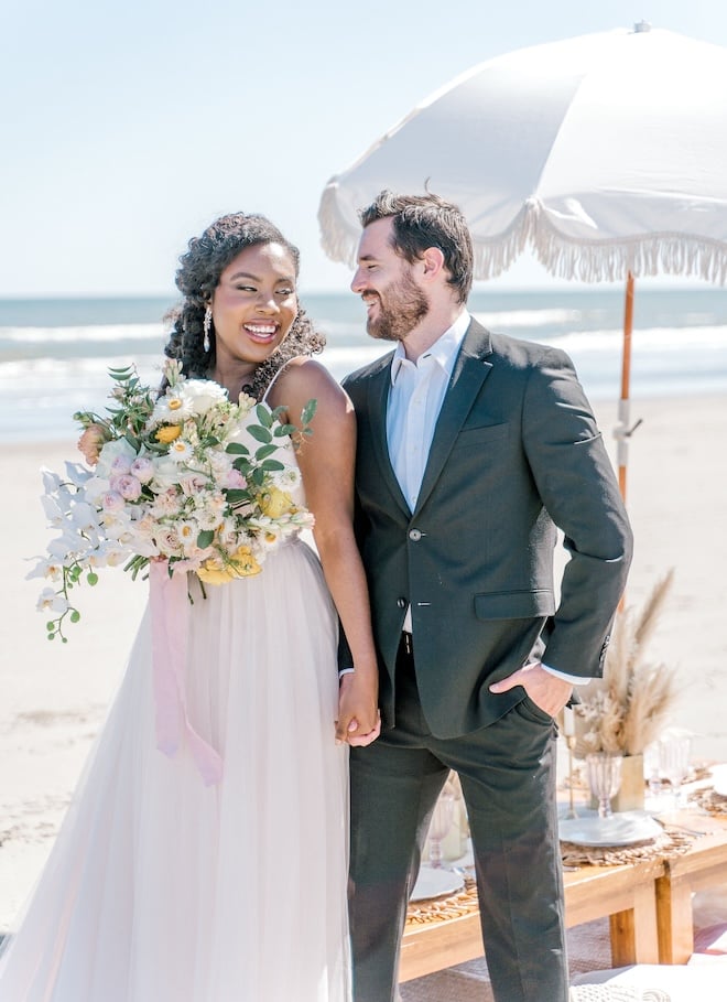 A bride and groom holding hands and smiling on a beach with a white umbrella behind them. 