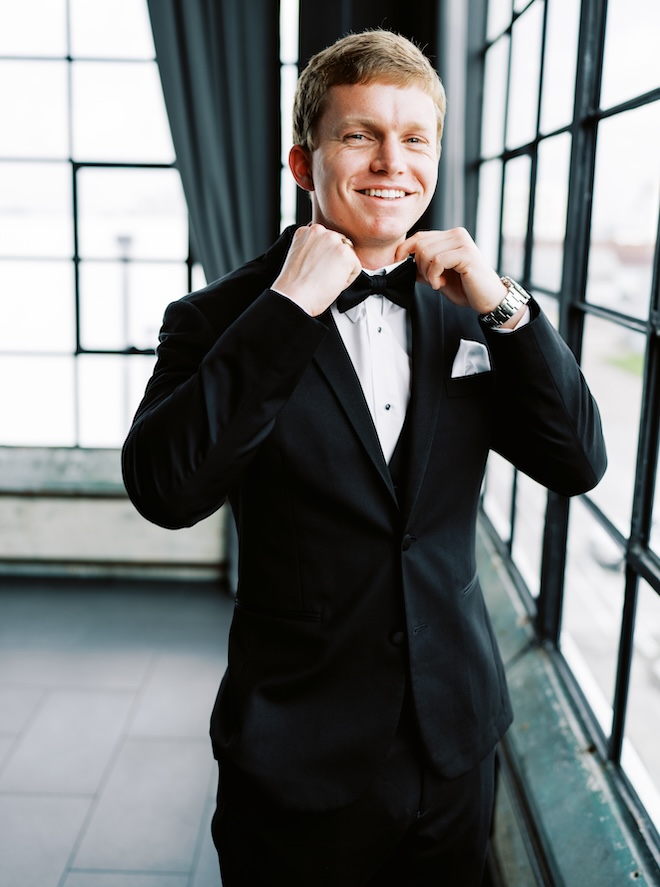 The groom adjusting his bow-tie while smiling at the camera. 