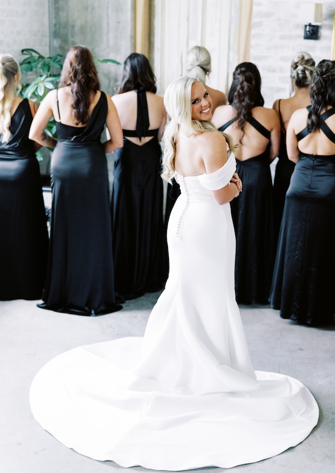 The bride smiling back at the camera with the bridesmaids wearing black before revealing her wedding gown. 