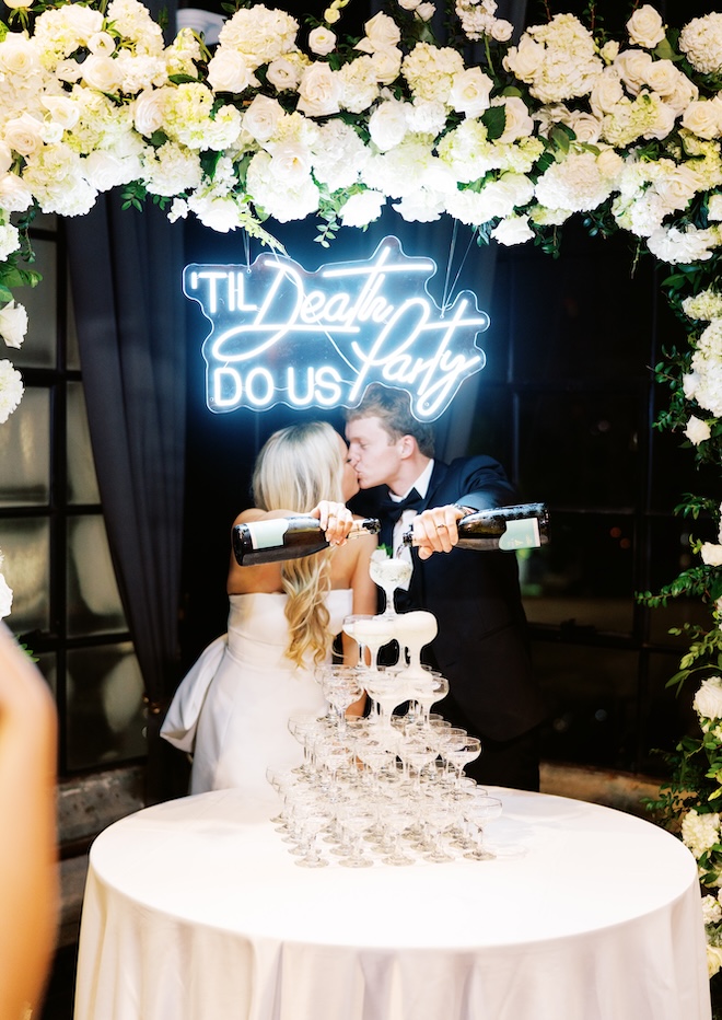 The bride and groom kissing and pouring champagne under a lit-up sign that says "'Til Death Do Us Party" above a champagne glass tower. 