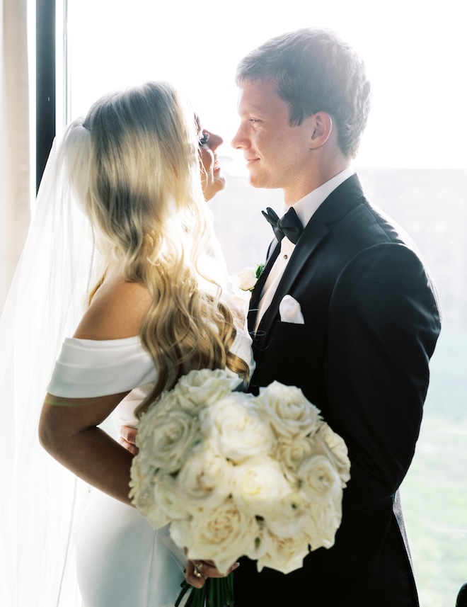 A bride wearing a veil and holding a large white bouquet smiling at the groom. 