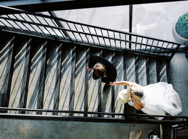 The groom holding the bride's hands and leading her up a black staircase. 