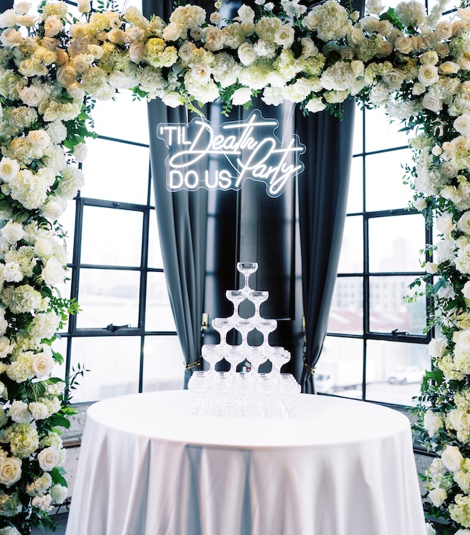 A lit-up sign that says "'Til Death Do Us Party" above a champagne glass tower. 