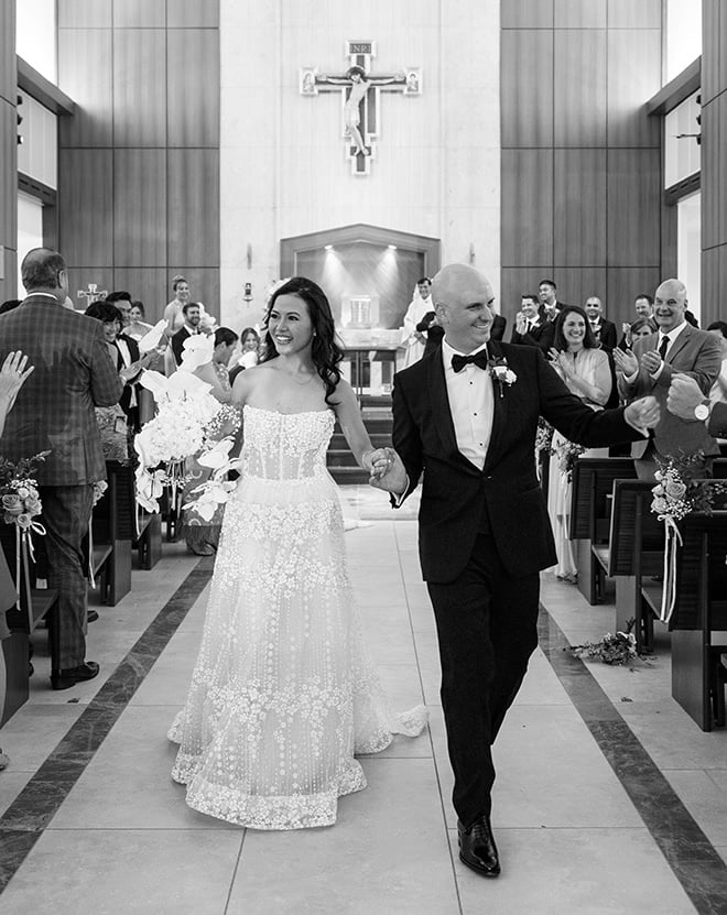 The bride and groom walk down the aisle at their wedding ceremony holding hands as guests cheer. 