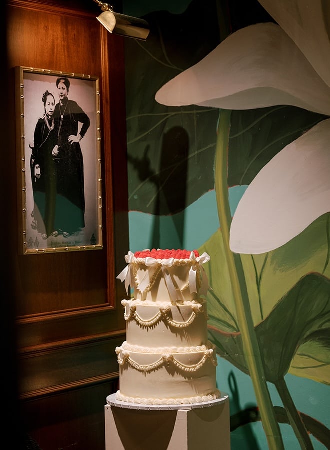 A three-tier white wedding cake is topped with white bows and red details. 