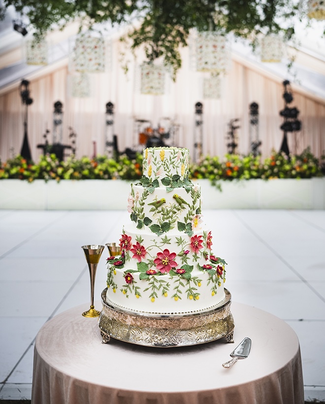 A four-tier colorful wedding cake with greenery, red and yellow florals and birds is the focal point of the wedding reception.