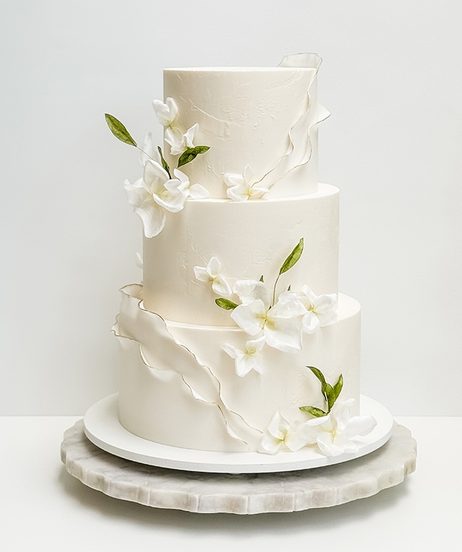 A three-tier white wedding cake with white florals and greenery designed by Lila Cake Shop.