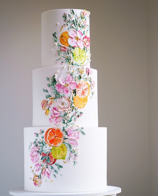 A three tier white wedding cake with hand painted colorful fruit and florals is featured in the new Weddings in Houston magazine. 