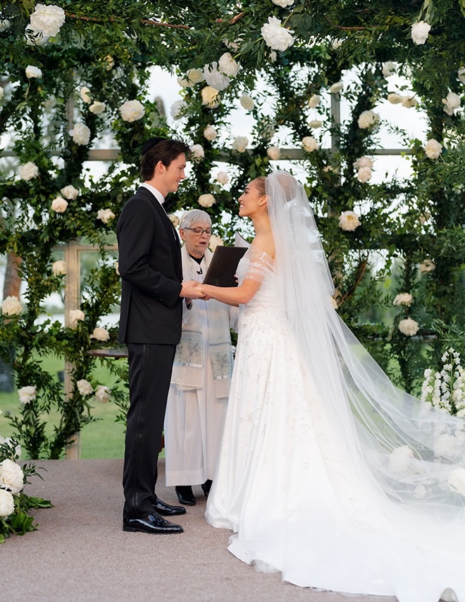 The couple hold hands at the altar with white florals and greenery as their backdrop.